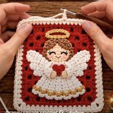 Red granny square with a white appliqued angel holding a red heart, gold halo and brown hair on wooden table