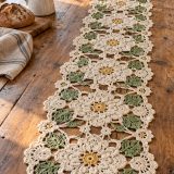 Cream floral crochet table runner with golden centers and sage leaves on a wooden table in sunlight