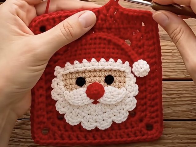 Red granny square with layered Santa face applique, white scalloped beard, red hat and pompom on wooden table