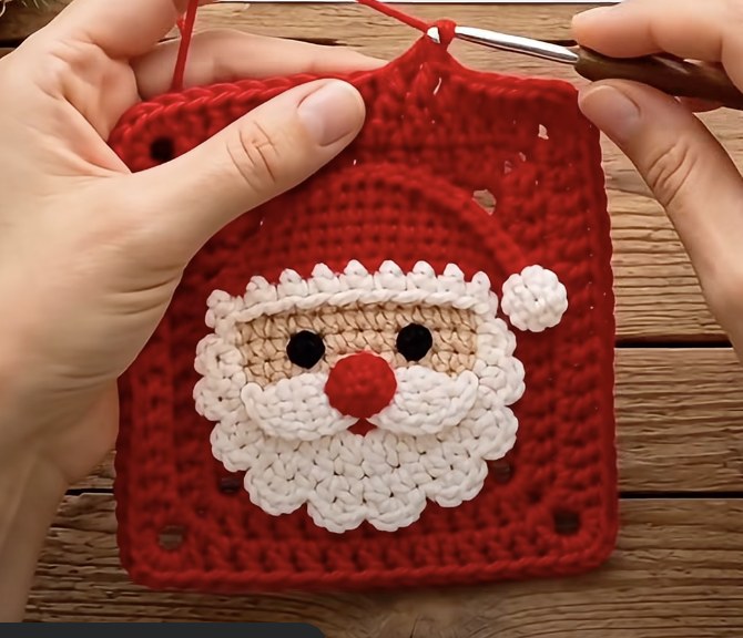 Red granny square with layered Santa face applique, white scalloped beard, red hat and pompom on wooden table